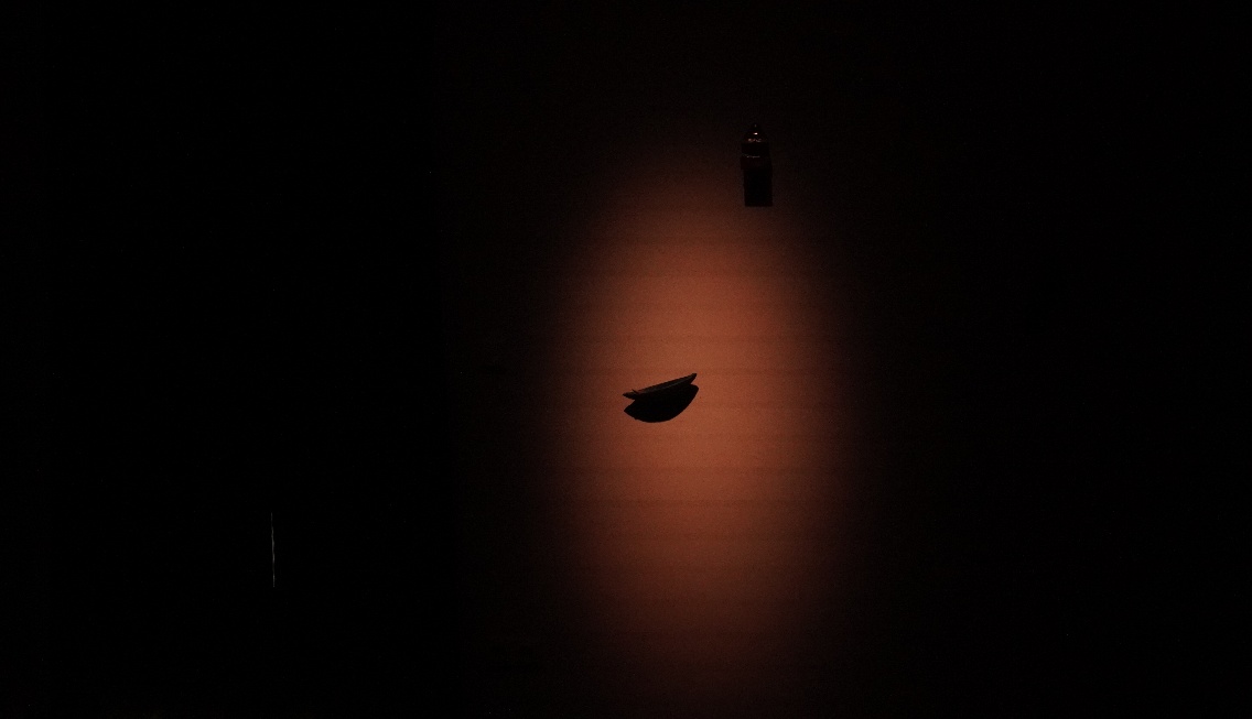 Light animation still — a single dark boat-like object mounted on a wall, illuminated by a concentrated warm orange glow in an otherwise black space.