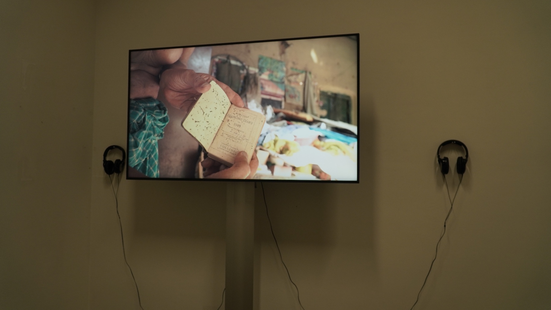 Exhibition view — a wall-mounted monitor showing a film still of hands carefully holding a small worn notebook, with headphones on either side for intimate viewing at Jan Van Eyck Academie.