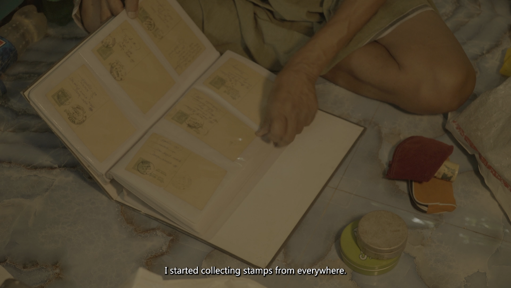 Film still — a cluster of old coins arranged on a colorfully painted decorative surface. Subtitle reads: 'Getting coins — I have even dreamt about it so many times — or buying books.'