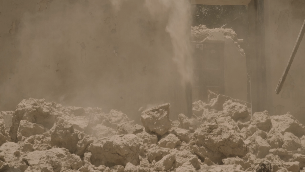 Film still — the demolition of a mud structure, dust billowing and rubble filling the frame, documenting the destruction of Selim's mud house.