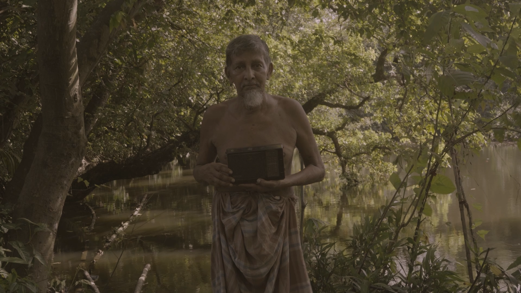 Film still — Khandakar Selim stands in a lush green forest, holding a dark object, reflecting on how trees are being cut down and on a future where forests may only be seen in museums.