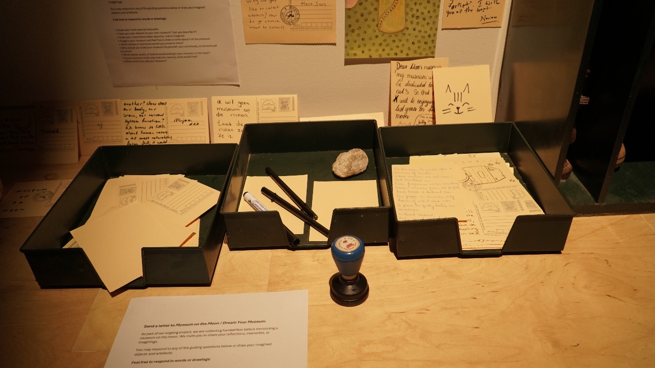 Interactive station — a desk with letter-writing materials: handwritten notes, postcards, a rubber stamp, and a printed invitation asking visitors to send a letter to 'Museum on the Moon / Dream Your Museum.'