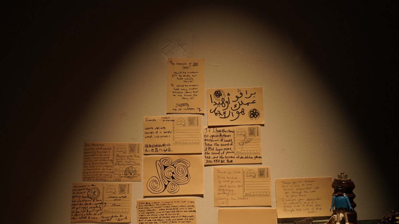 Interactive station — letters and postcards written by visitors, pinned to the wall under a single spotlight, alongside a small toy figure. These letters will travel to Kelepara village, India, to be held in the future community museum.