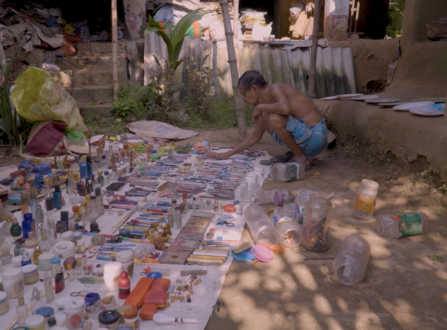 Film still from Museum on the Moon: Khandakar Selim crouches over his collection spread across the ground outside his mud house — hundreds of objects laid out in rows, including bottles, pens, tools, containers, and miscellaneous artifacts accumulated over five decades.