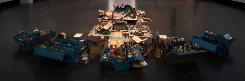 Full panoramic view of the Dream Your Museum installation at Akademie der Kunste, Berlin, 2022. All blue metal trunks are open and arranged symmetrically around a central spread of Khandakar Selim's collected objects displayed on the gallery floor. Photo credit: Dotgain.info, Berlin Biennale.