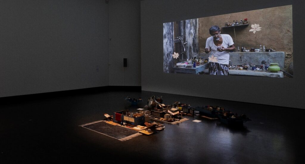 Installation view of Dream Your Museum at Akademie der Kunste, Berlin, 2022. The film is projected on the wall — showing Khandakar Selim at work in his home, surrounded by his accumulated objects — while the installation of trunks and collected items is displayed on the floor below. Photo credit: Dotgain.info, Berlin Biennale.