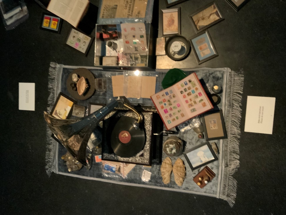 Detail view of the Dream Your Museum installation — overhead close-up of objects displayed on a mat, including a gramophone record, stamp collection albums, coins, small framed prints, and other accumulated items from Khandakar Selim's domestic museum.
