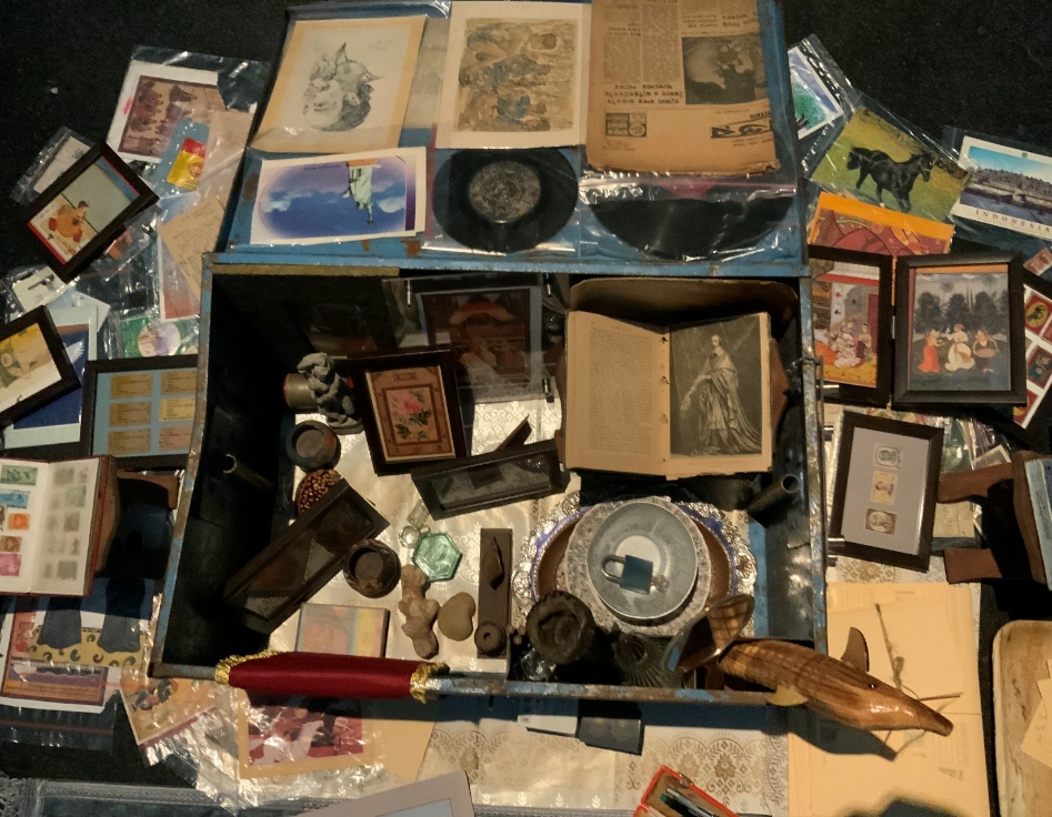Detail view of an open blue metal trunk in the Dream Your Museum installation, revealing a dense arrangement of eclectic objects: old photographs, printed images, small bottles, books, a vinyl record, a horse figurine, coins, stamps, and a large red pen — the contents of Selim's domestic archive.