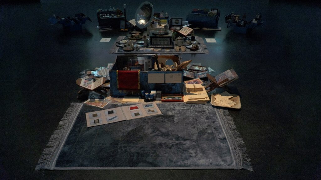 Overhead installation view of Dream Your Museum at Akademie der Kunste, Berlin, 2022. The darkened gallery space shows the full arrangement of blue metal trunks and collected objects spread across sitting mats on the floor, with the film projection visible at the far end.