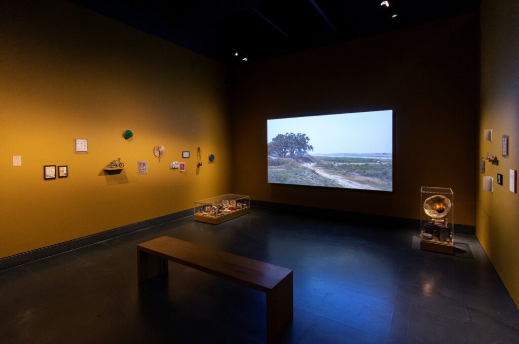 Installation view of Dream Your Museum at the Jameel Prize 7 Exhibition, Victoria and Albert Museum, London, 2024. The work is installed in a warm amber-walled gallery: the film is projected on the rear wall showing a rural landscape with a tree and a dirt road, while framed objects and small works are mounted on the side walls and display cases are arranged on the gallery floor.