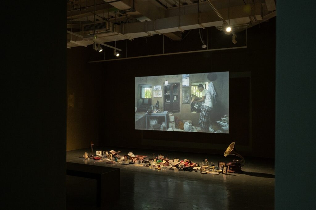 Installation view of Dream Your Museum at Hayy Jameel, Jeddah, Saudi Arabia. The darkened gallery features the film projected on the rear wall — a scene of Khandakar Selim in his home surrounded by accumulated objects — with the metal trunks and collected items arranged on a raised platform in the foreground, and a gramophone visible to the right.