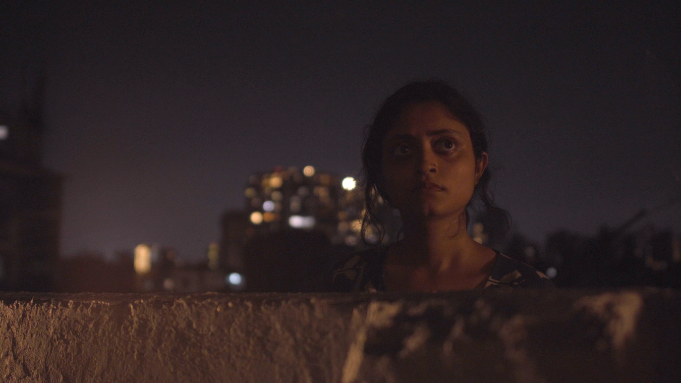 Film still from A Collision in the Wind, 2024. Close-up of a young woman at night, her face illuminated against the city skyline behind her, looking directly into the camera — a moment of raw, still grief amid the urban environment.