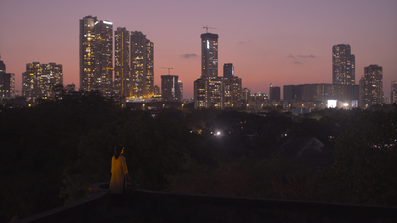 Film still from A Collision in the Wind, 2024. A lone figure in yellow stands on a rooftop or elevated ground in silhouette against a dusky pink-and-purple city skyline at dusk, high-rise buildings lit in the background — a scene of solitude and longing.