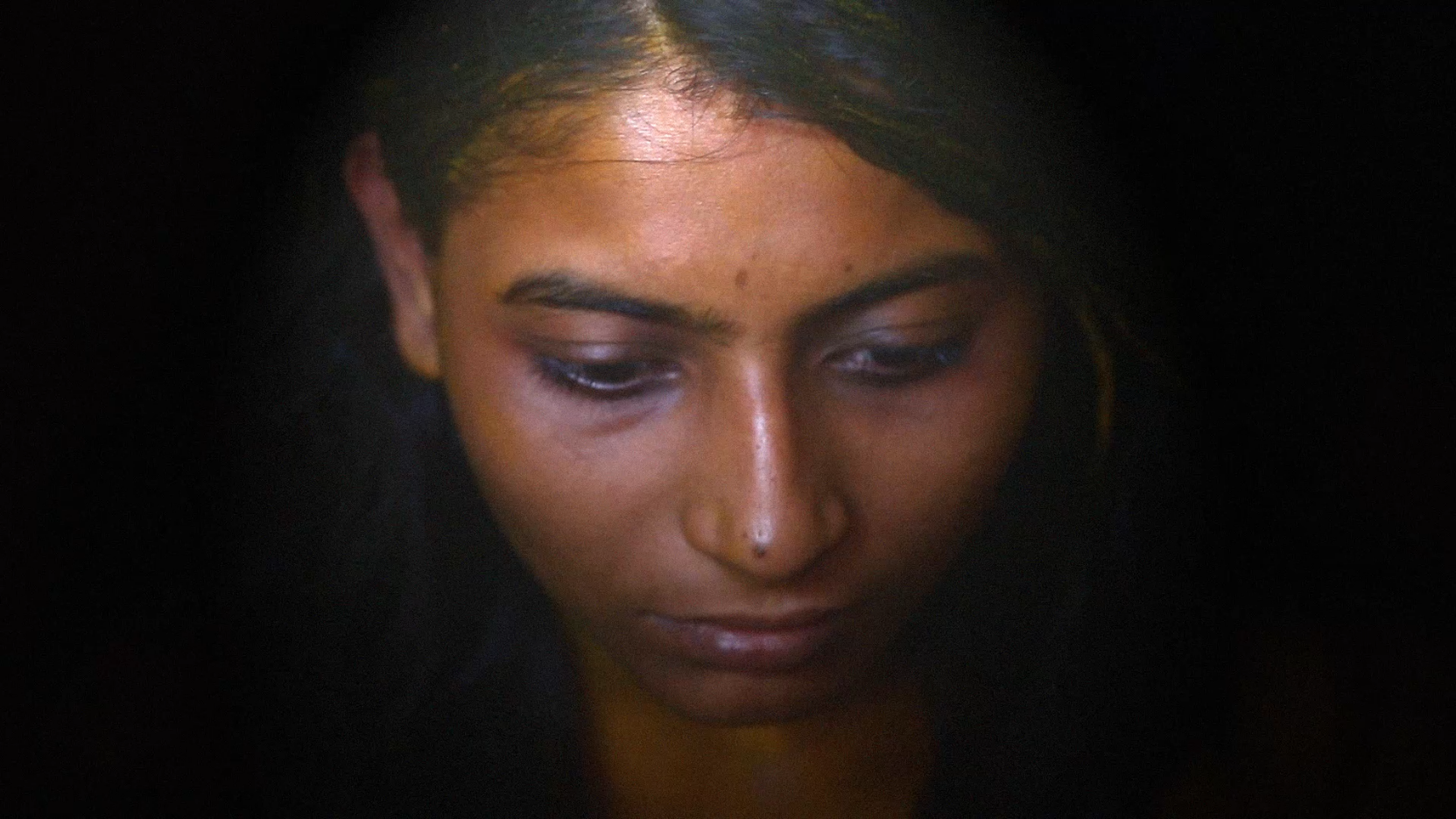 Video still from Beyond Monochrome — a close-up portrait of a woman’s face, partially illuminated against a deep black background. Her downward gaze conveys interiority and quiet resistance. Video art, 4 min 11 sec, 2019.