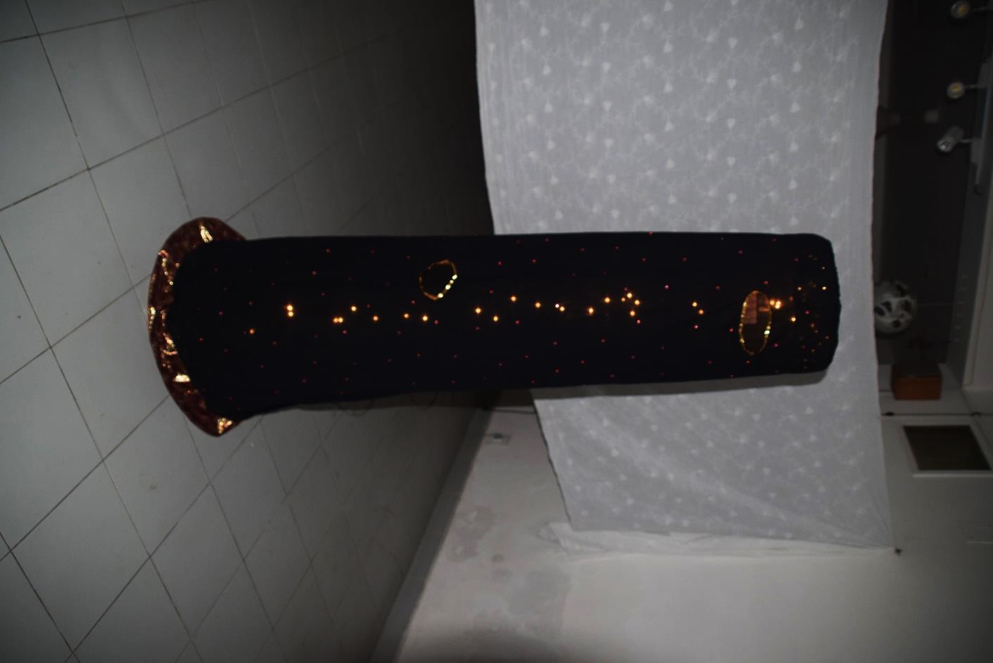 The Home and the Globe, installation view, 2018. A tall, tapering cylindrical structure of black cloth adorned with embroidery stones and illuminated by a chain of zero-watt lights glowing through pinprick perforations. Two circular mirror-framed eyeholes are visible along the surface, inviting viewers to peer inside. The piece stands approximately 66 inches high against a white tiled wall.