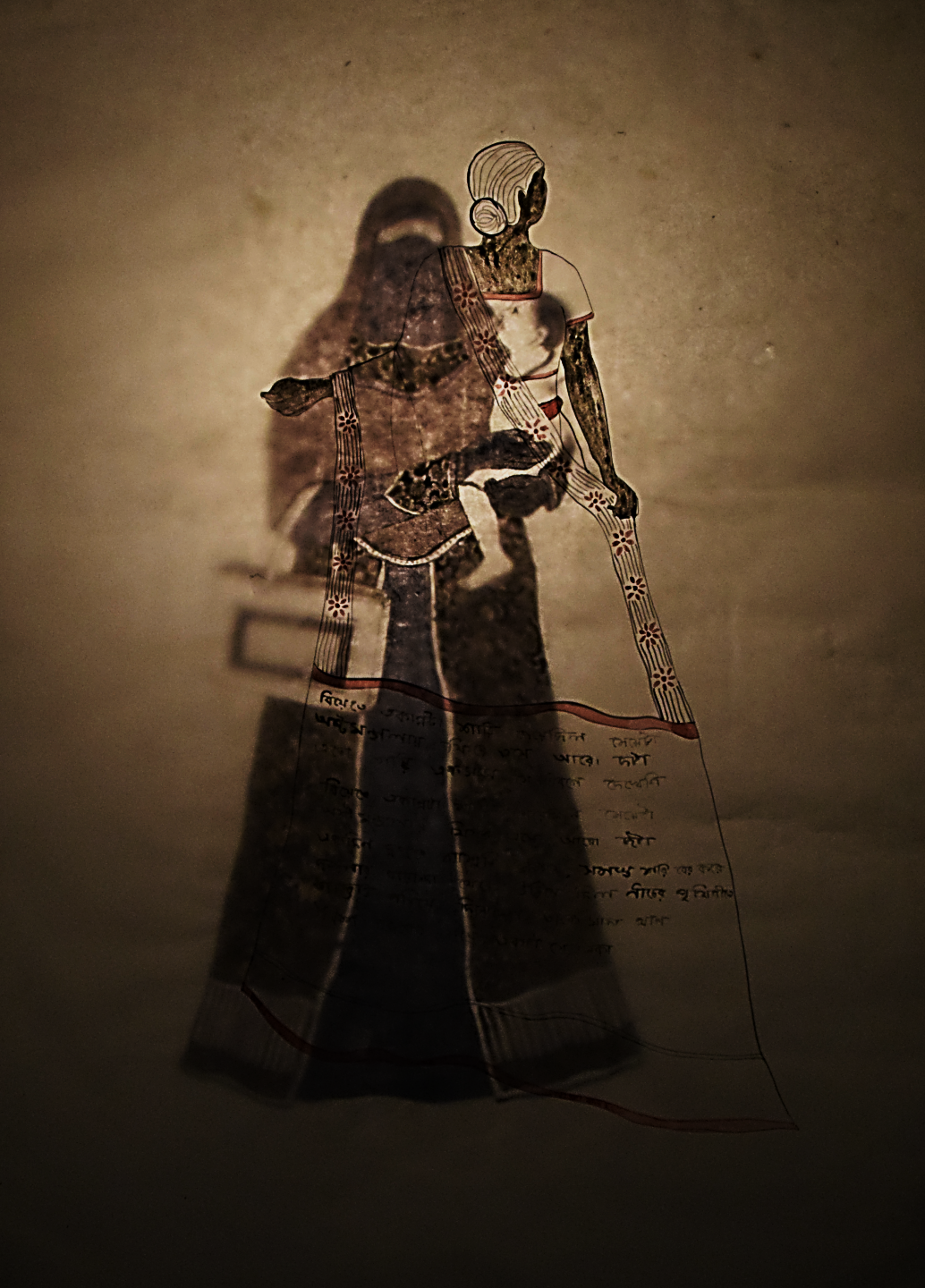 Burden of Sorrow, drawing installation, two drawings on paper with tungsten bulb backlight, 24 x 18 inches, 2018. Photographed in situ with backlit illumination, the layered drawings merge into a single glowing image: a fully veiled woman in an ornate dark burqa holds a small child and a long ribbon or scroll bearing Bengali script, while a second drawn layer creates a ghostly double exposure. The warm amber light of the tungsten bulb suffuses the composition.