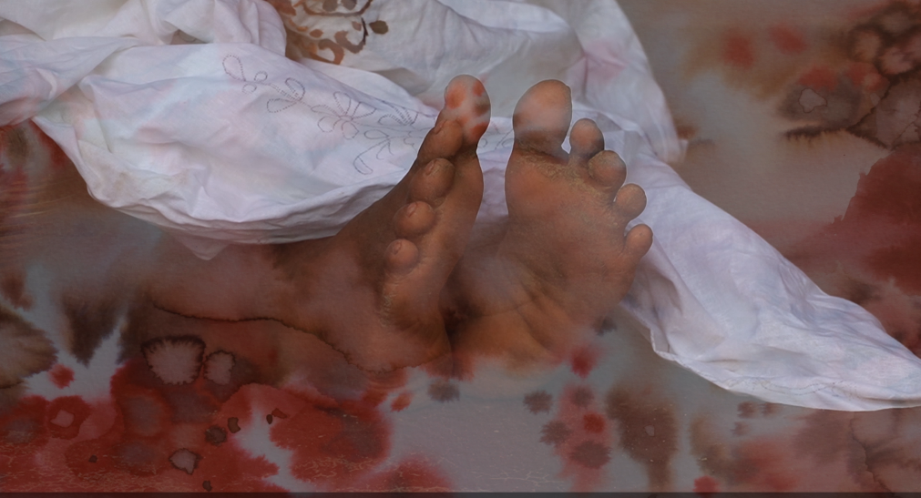 Still from The Last Dream I Saw — bare feet surrounded by red paint or liquid bleeding across a white cloth, evoking themes of trauma and violence.