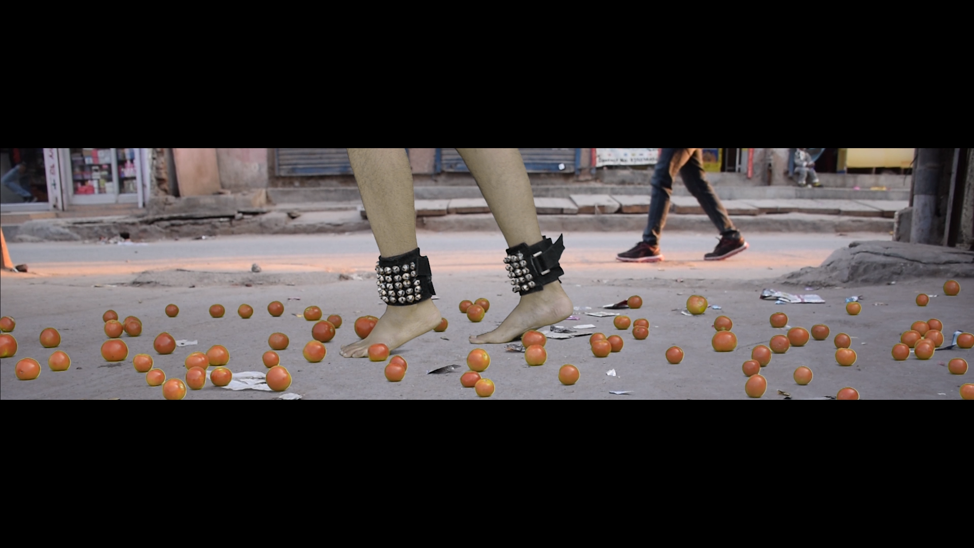 Video still from Ungraceful Melody — a woman's bare feet wearing studded black leather ankle cuffs stand among scattered tomatoes on a public street, passersby visible in the background.
