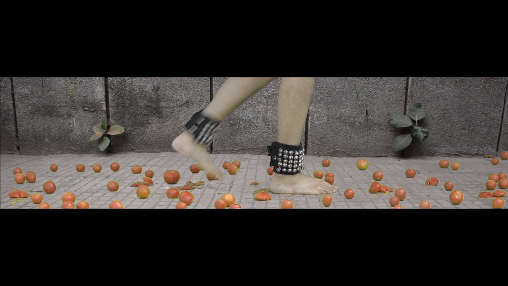 Video still from Ungraceful Melody — a woman's foot in a studded black ankle cuff steps among scattered tomatoes against a concrete wall, referencing both freedom and confinement.