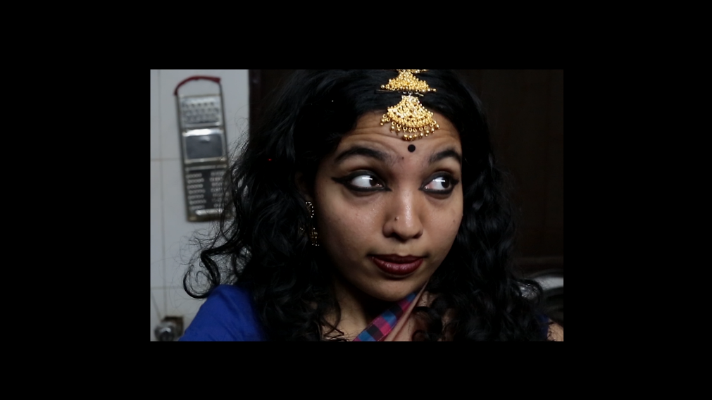 Video still from Ungraceful Melody — a close portrait of a woman wearing traditional Indian bridal jewelry and blue attire, looking directly at the viewer with an ambiguous expression.
