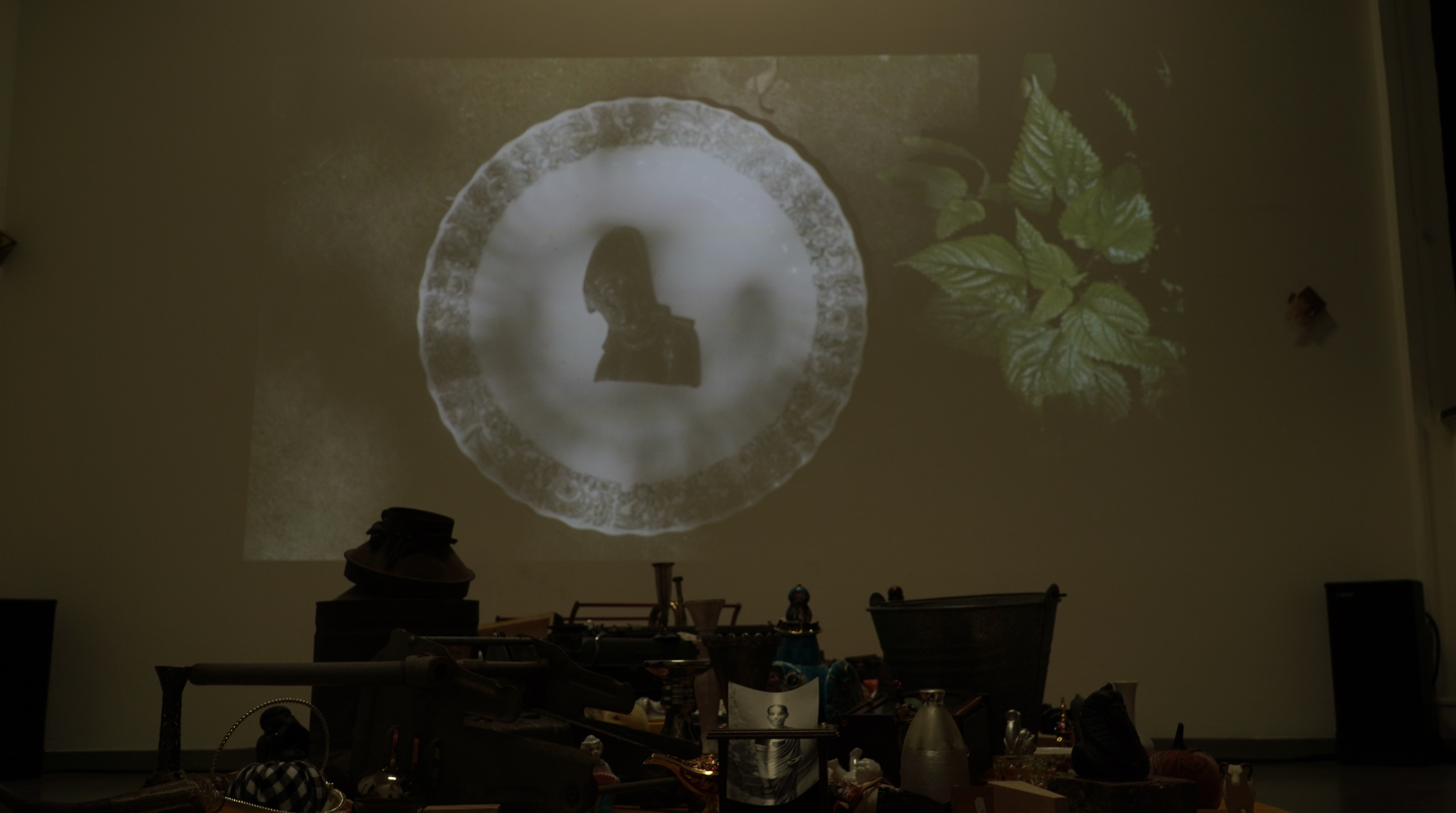 Film projection on the wall at Jan Van Eyck Academie — a silhouetted figure appears inside a projected decorative plate, alongside botanical imagery, above the table of collected objects below.