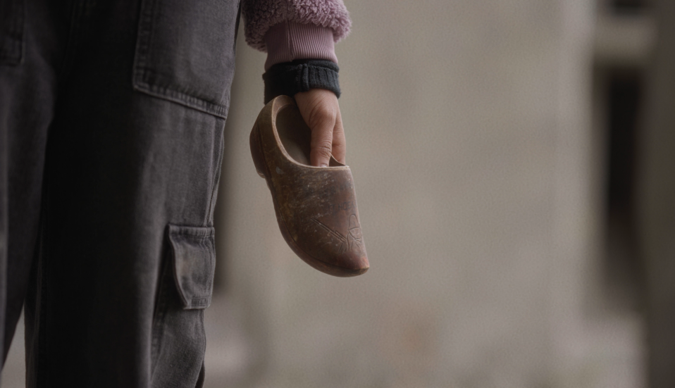Film still — a hand holding a Dutch wooden clog, one of the collected objects gathered from the Netherlands for the archive.