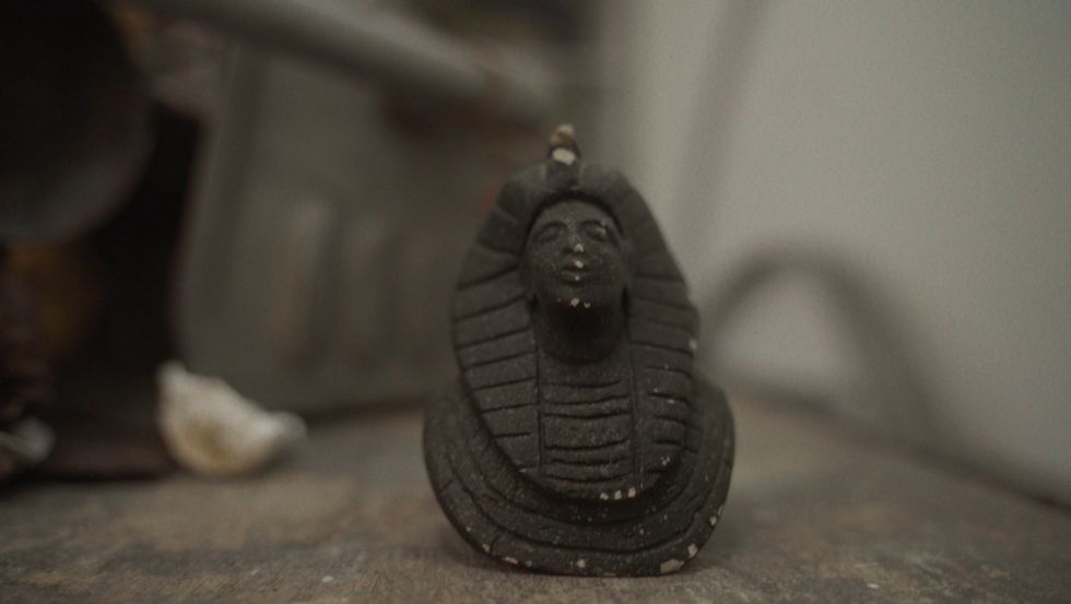 Film still — a small Egyptian pharaoh figurine resting on a surface, among the eclectic found objects collected across the Netherlands.