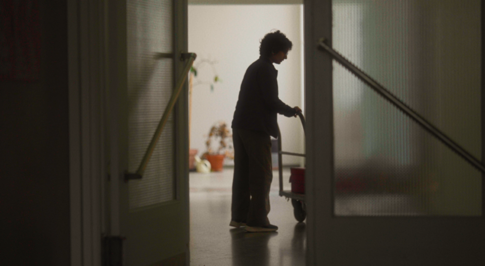 Film still — an elderly figure silhouetted in a doorway, evoking themes of thresholds, memory, and the passage of time.