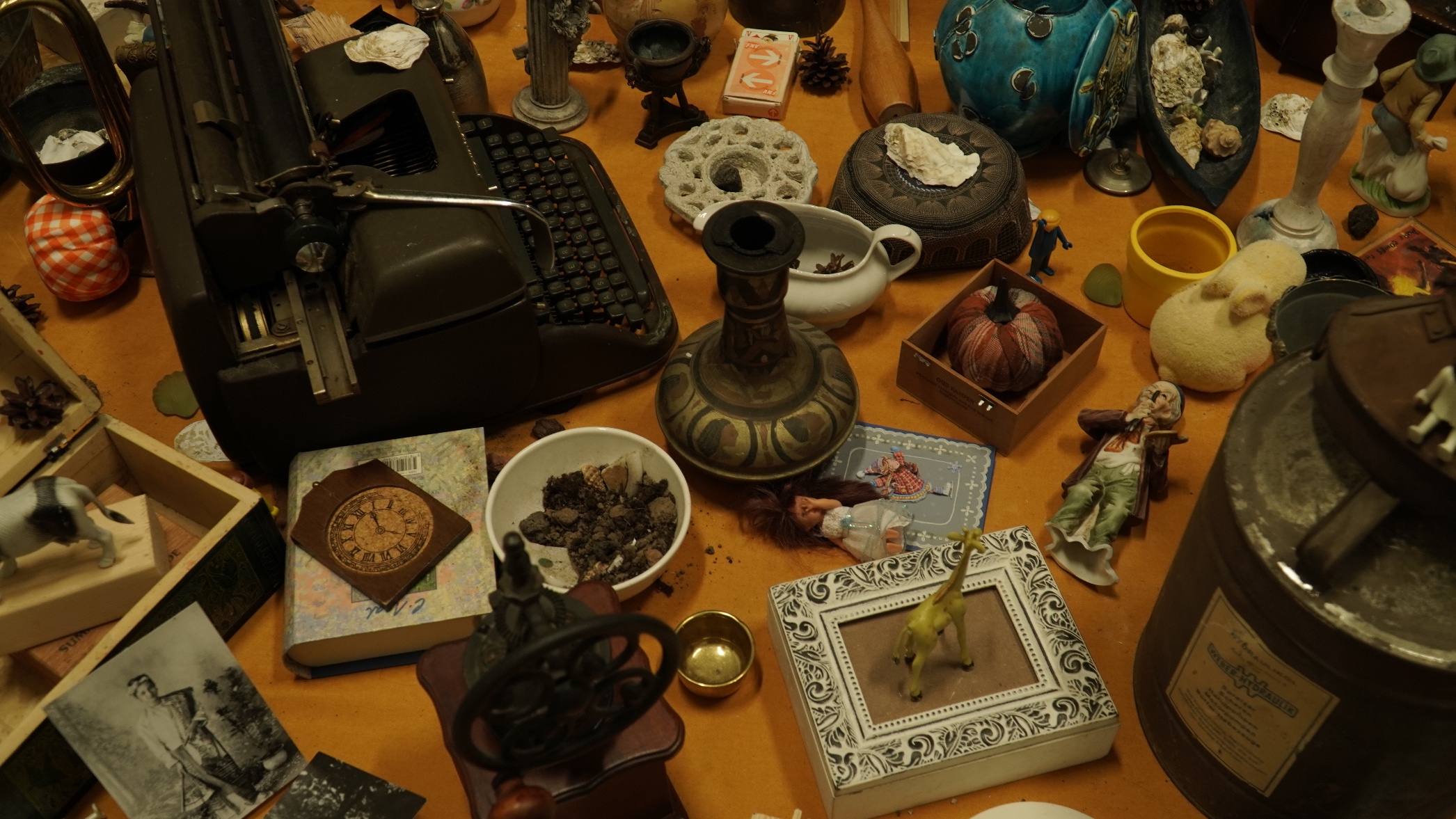 Detail of collected objects — a close-up of the table's surface revealing a typewriter, ornate vases, a clock face, ceramic figurines, a toy cow, and other gathered materials.