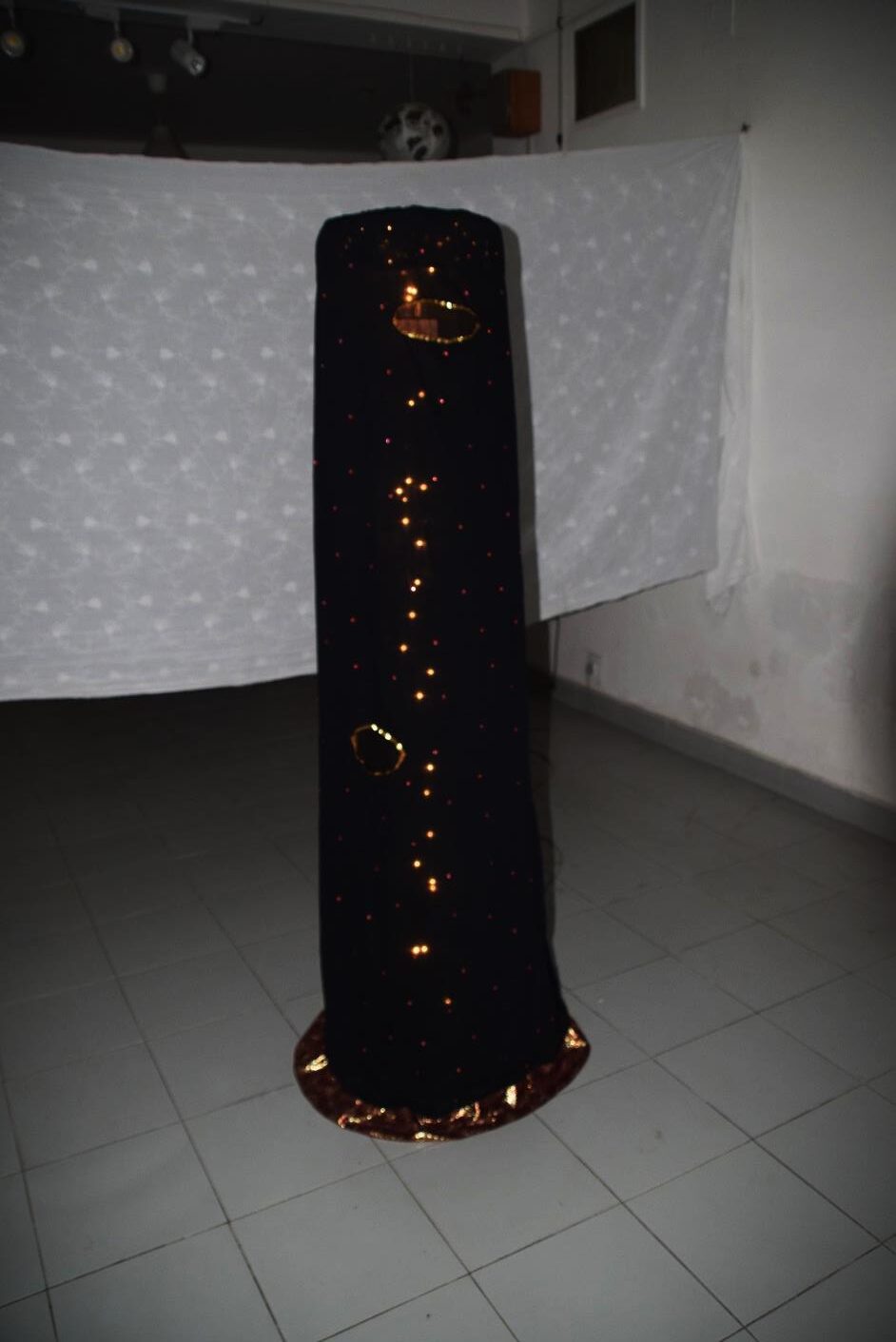 The Home and the Globe, installation view, 2018. A tall, tapering cylindrical structure of black cloth adorned with embroidery stones and illuminated by a chain of zero-watt lights glowing through pinprick perforations. Two circular mirror-framed eyeholes are visible along the surface, inviting viewers to peer inside.